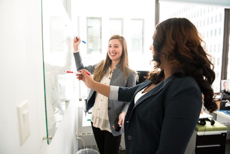 Dwie młode kobiety w biurze śmiejące się do siebie przed tablicą na ścianie do zapisywania notatek